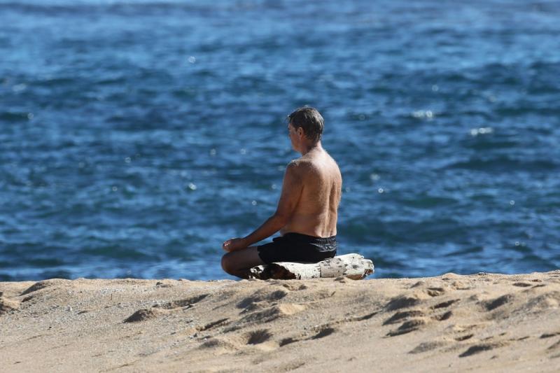 Pierce Brosnan, complet ud, în poziția lotus, meditând în fața oceanului