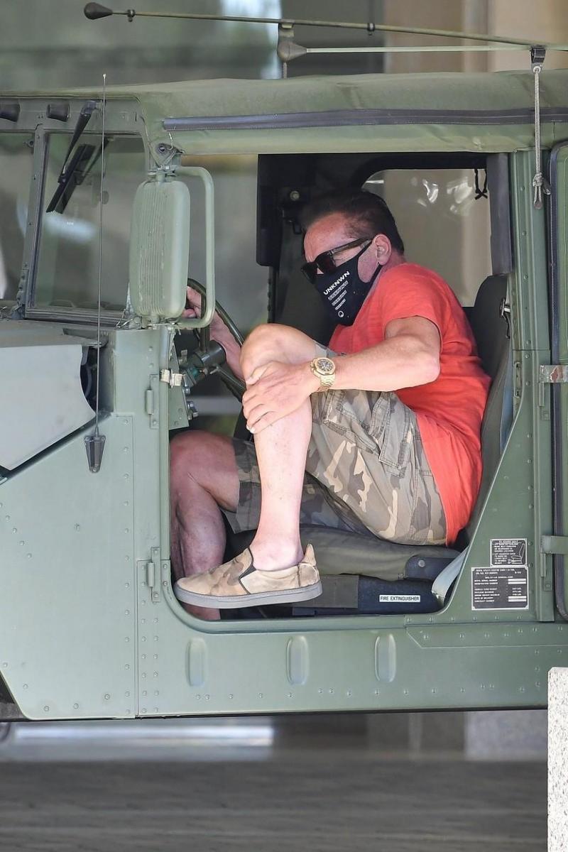 Arnold Schwarzenegger, fotografiat de paparazzi la bordul unui Hummer. Actrorul poartă mască de protecție și este îmbrăcat într-un tricou roșu și pantaloni scur