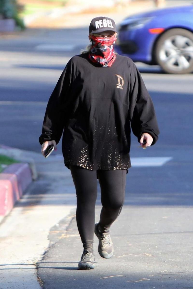 Rebel Wilson, îmbrăcată într-o ținută închisă la culoare, cu ochelari, șapcă și mască de protecție