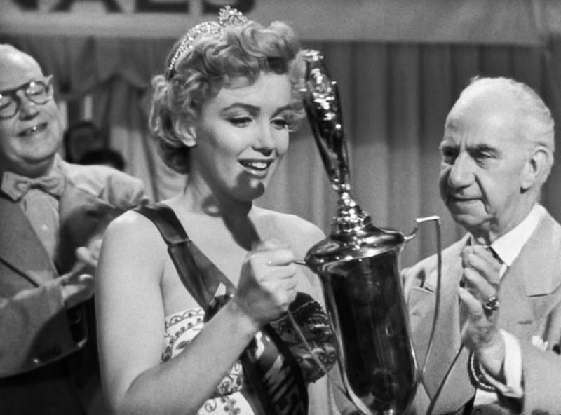 Marilyn Monroe înconjurată de barbati, tinand un premiu in maini