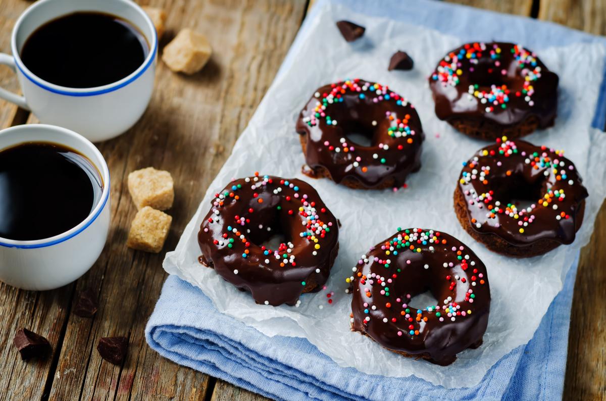 Cinci gogoși cu ciocolată, așezate pe un șervet de bucătărie, alături de două cani în care se află cafea.