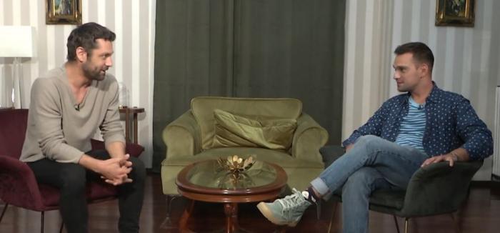 Alecsandru Dunaev poartă pantaloni de culoare închisă și o bluză gri
