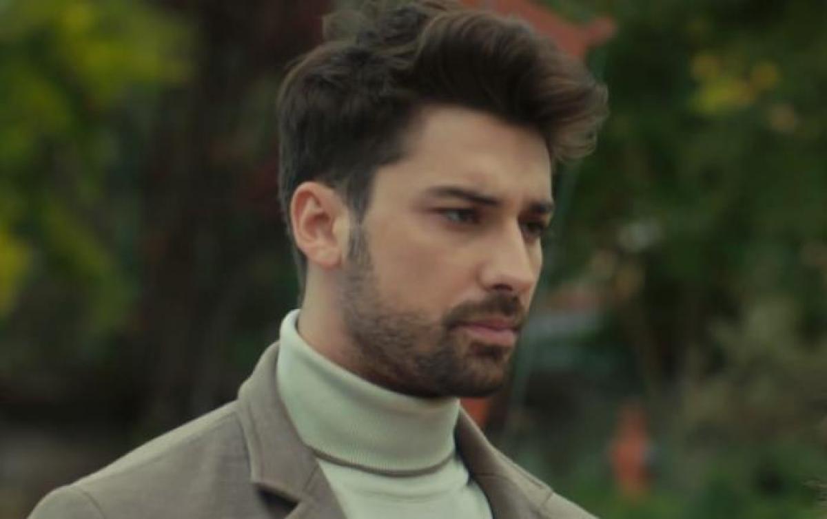 Alp Navruz poartă o maletă și un palton gri