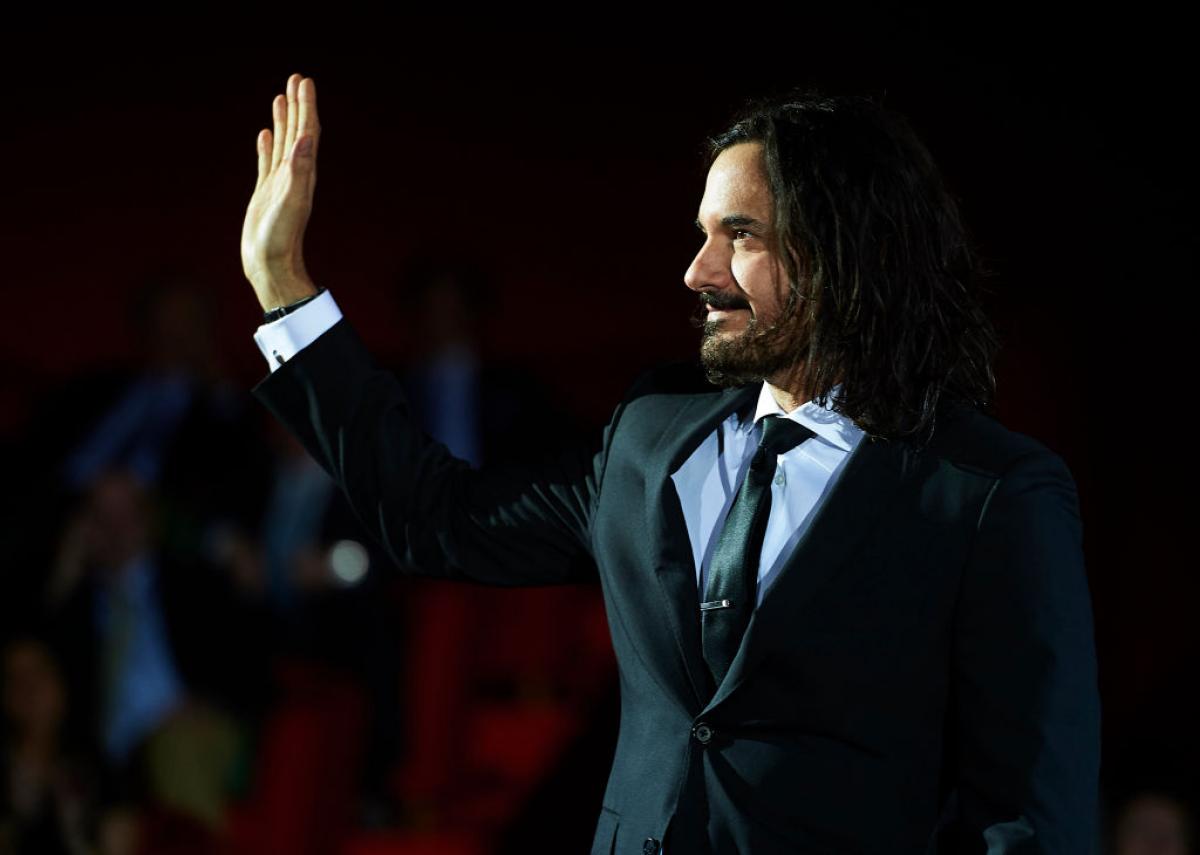 Mario Cimarro poartă o cămașă albă, cravată și un sacou închis la culoare. Are părul desprins, cârlionțat