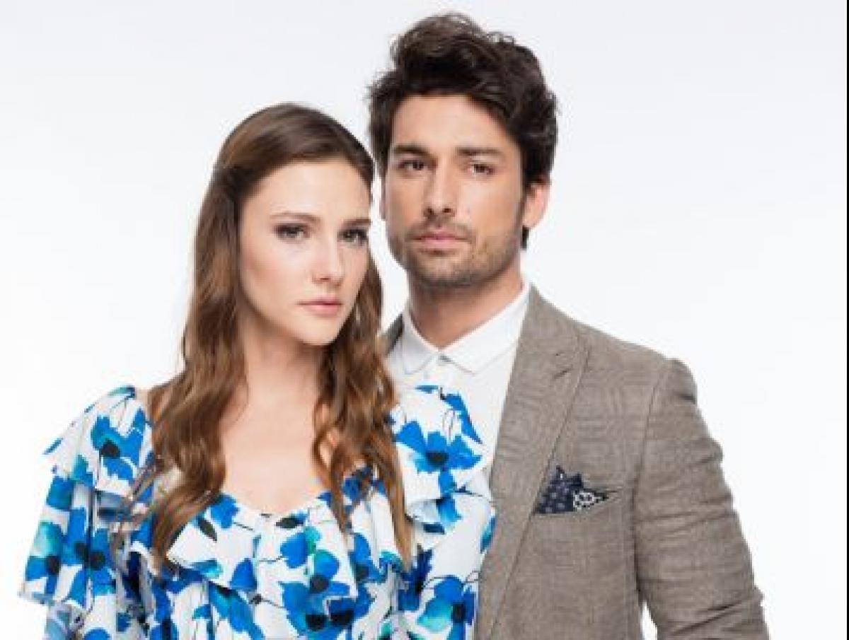 Alp Navruz poartă un sacou gri și stă alături de partenera lui din serial