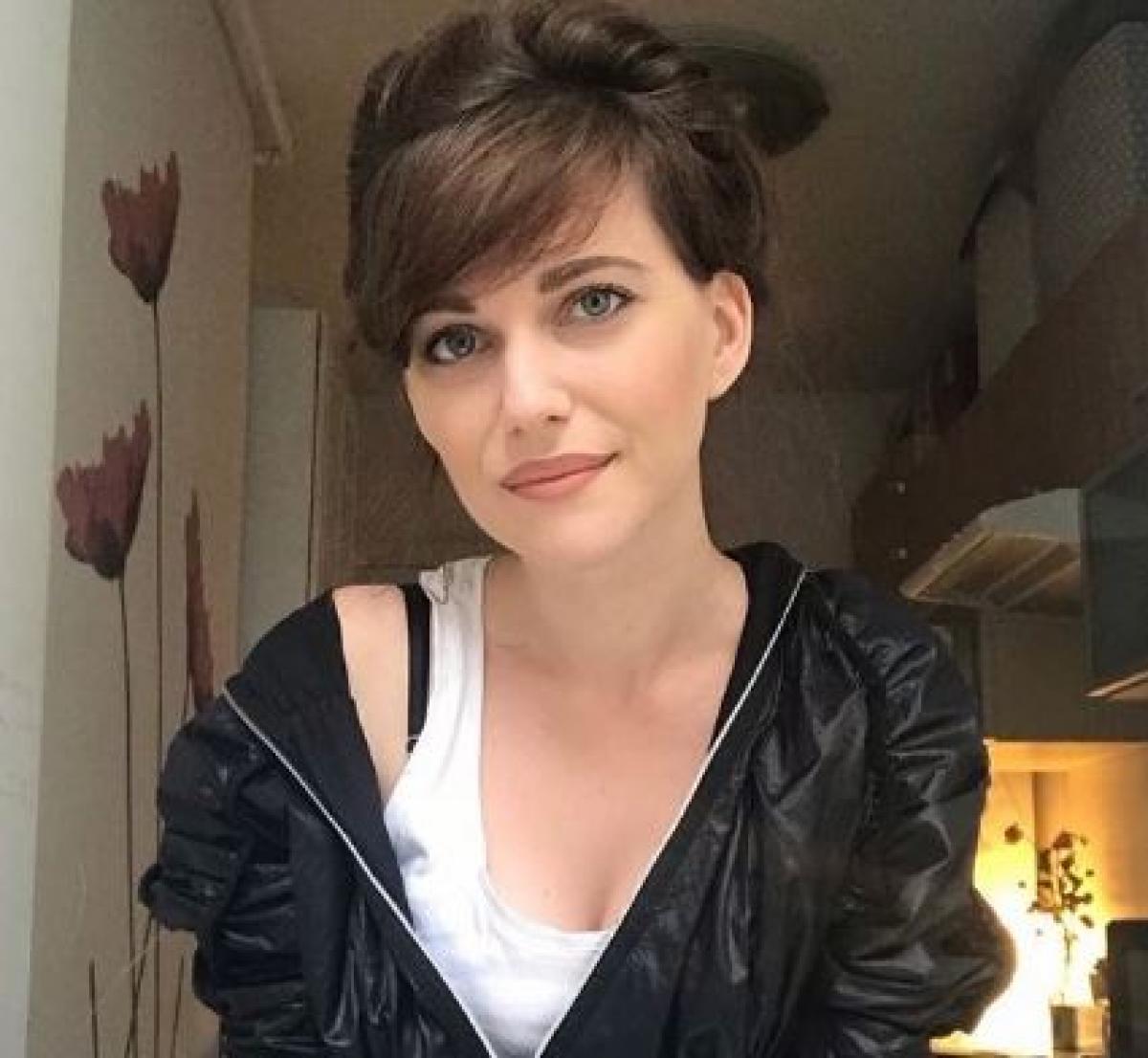 Alexandra Gherman este îmbrăcată într-un maiou alb și o geacă neagră care îi cade lejer pe umăr. Jurnalista are părul prins și breton