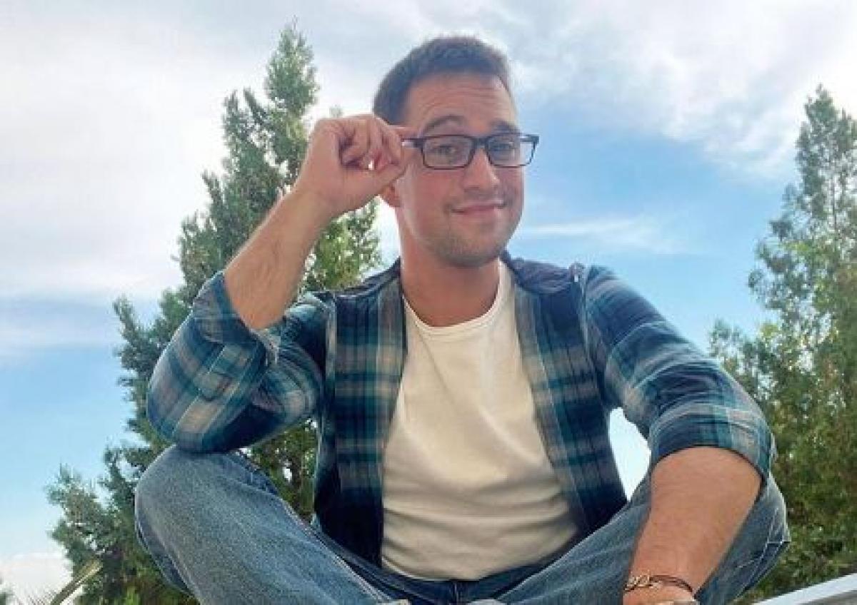 Vlad Gherman are un tricou alb și o cămașă albastră cu dungi. Actorul poartă ochelari