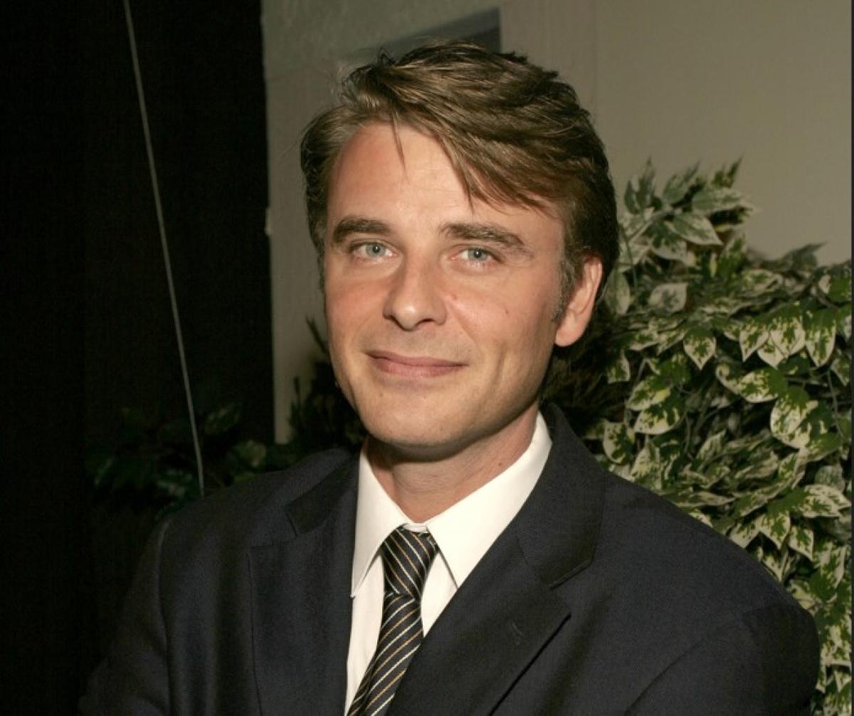 Juan Pablo Gamboa poartă un costum închis la culoare și are o cravată albă