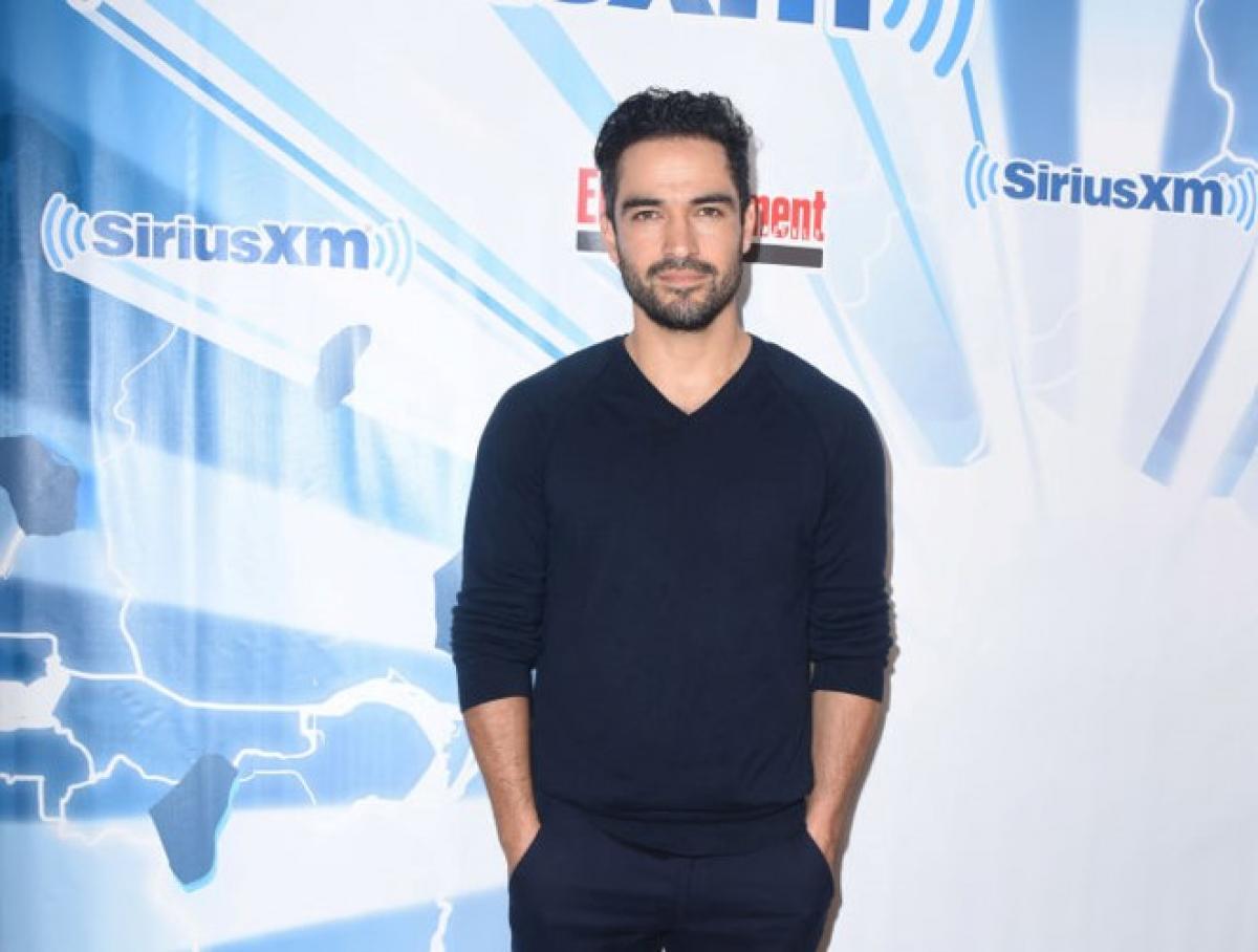 Alfonso Herrera poartă pantaloni închiși la culoare și o maletă de aceeași culoare. Artistul își ține mâinile în buzunare