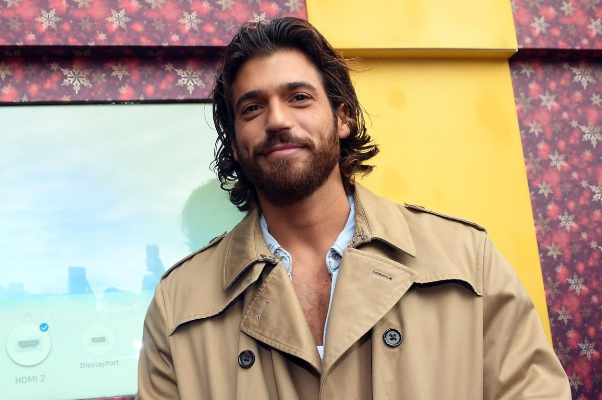 Can Yaman poartă o cămașă deschisă la gât și are un palton maro deschis