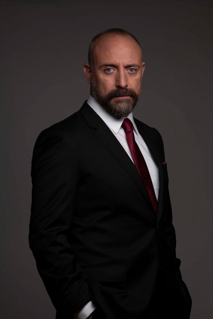 Actorul Halit Ergenç îmbrăcat în costum negru, fotografiat pe fundal gri