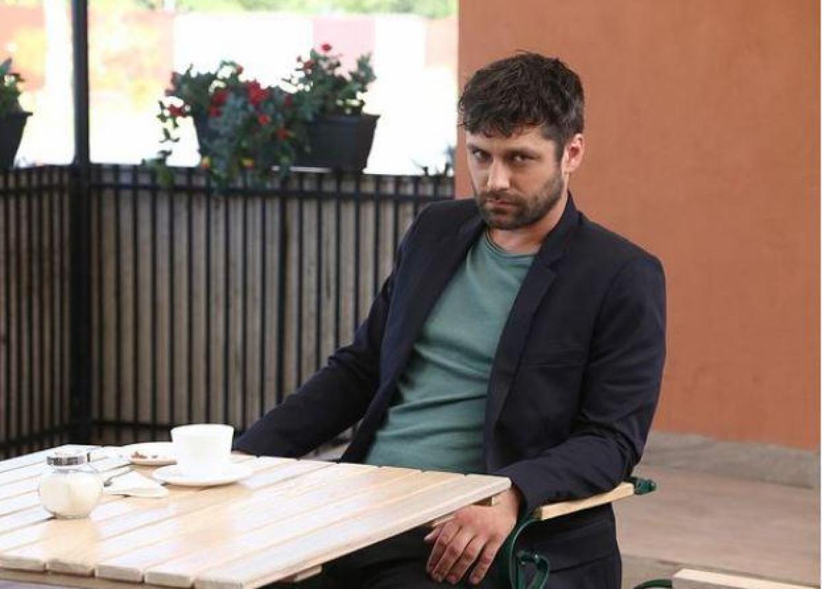 Alecsandru Dunaev, îmbrăcat cu un sacou, privește drept în obiectivul camerei de fotografiat, în timp ce stă pe scaun, la o masă.