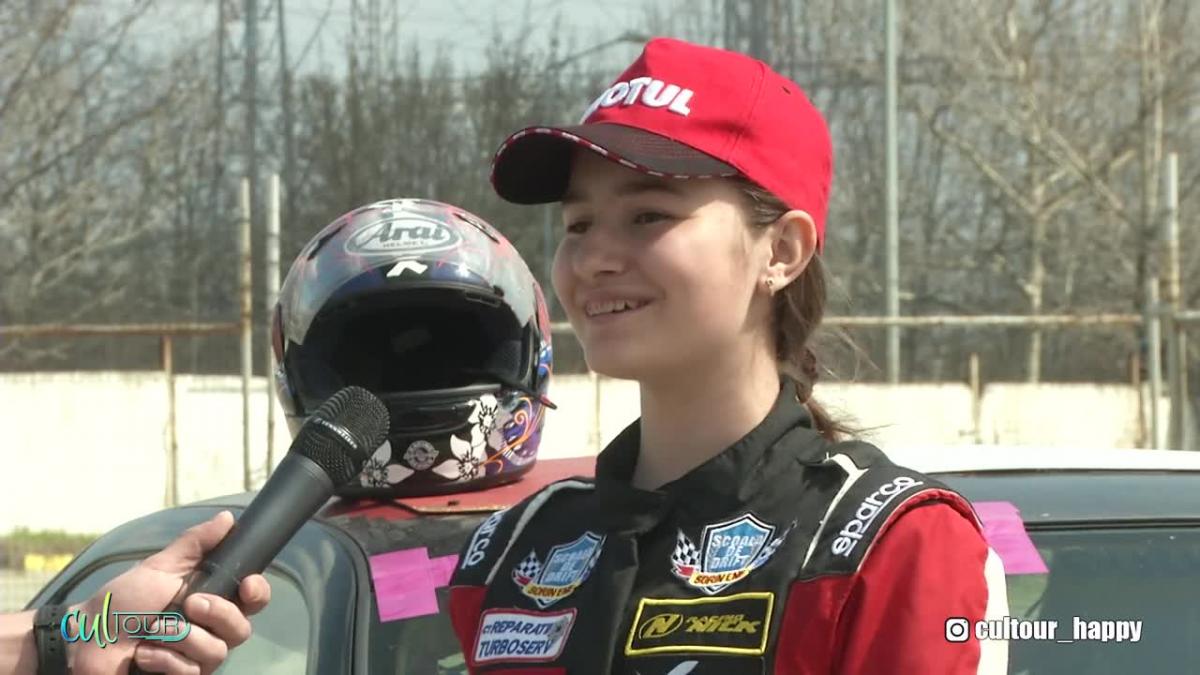 Alice Ene, în echipament de pilot de curse, cu șapcă roși pe cap, acordă un interviu reporterului Happy Channel.