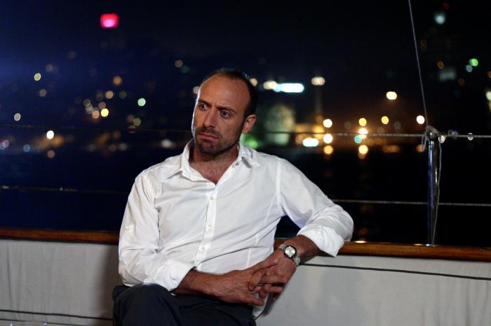 Halit Ergenç poartă o cămașă albă și pantaloni închiși la culoare