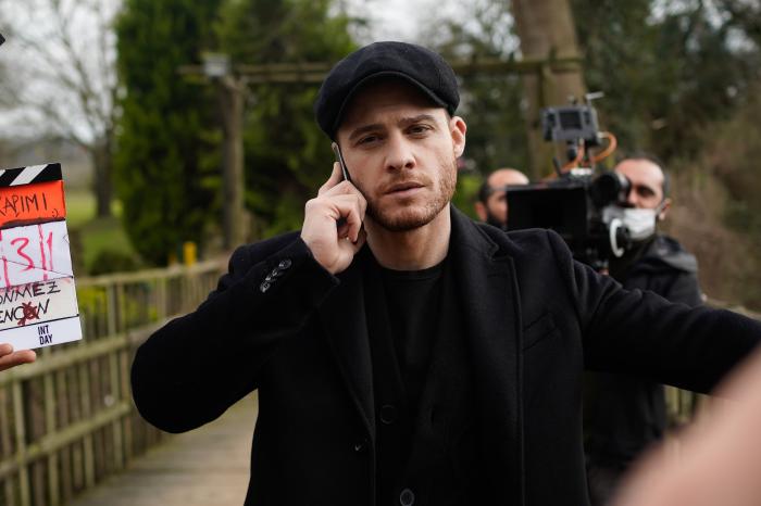 Kerem Bursin este îmbrăcat în negru, vorbește la telefon și are pe cap o beretă neagră