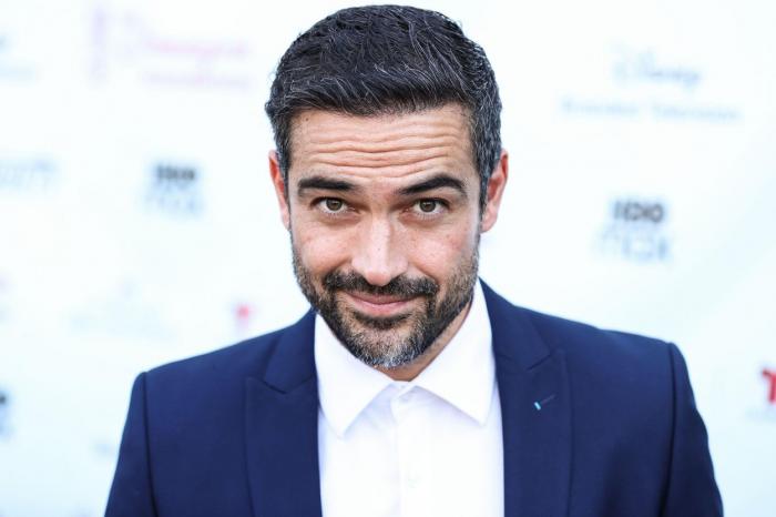 Alfonso Herrera poartă o cămașă albă, fără cravată sau papion, se strâmbă și poartă un sacou albastru închis