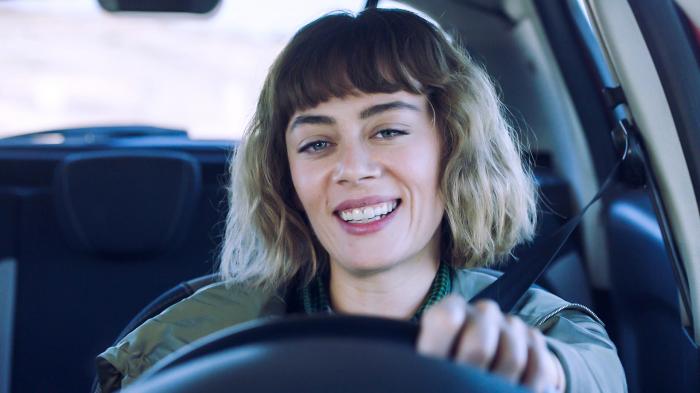 Demet Evgar se află la volanul unei mașimi, zâmbește și ține mâna stângă pe volan