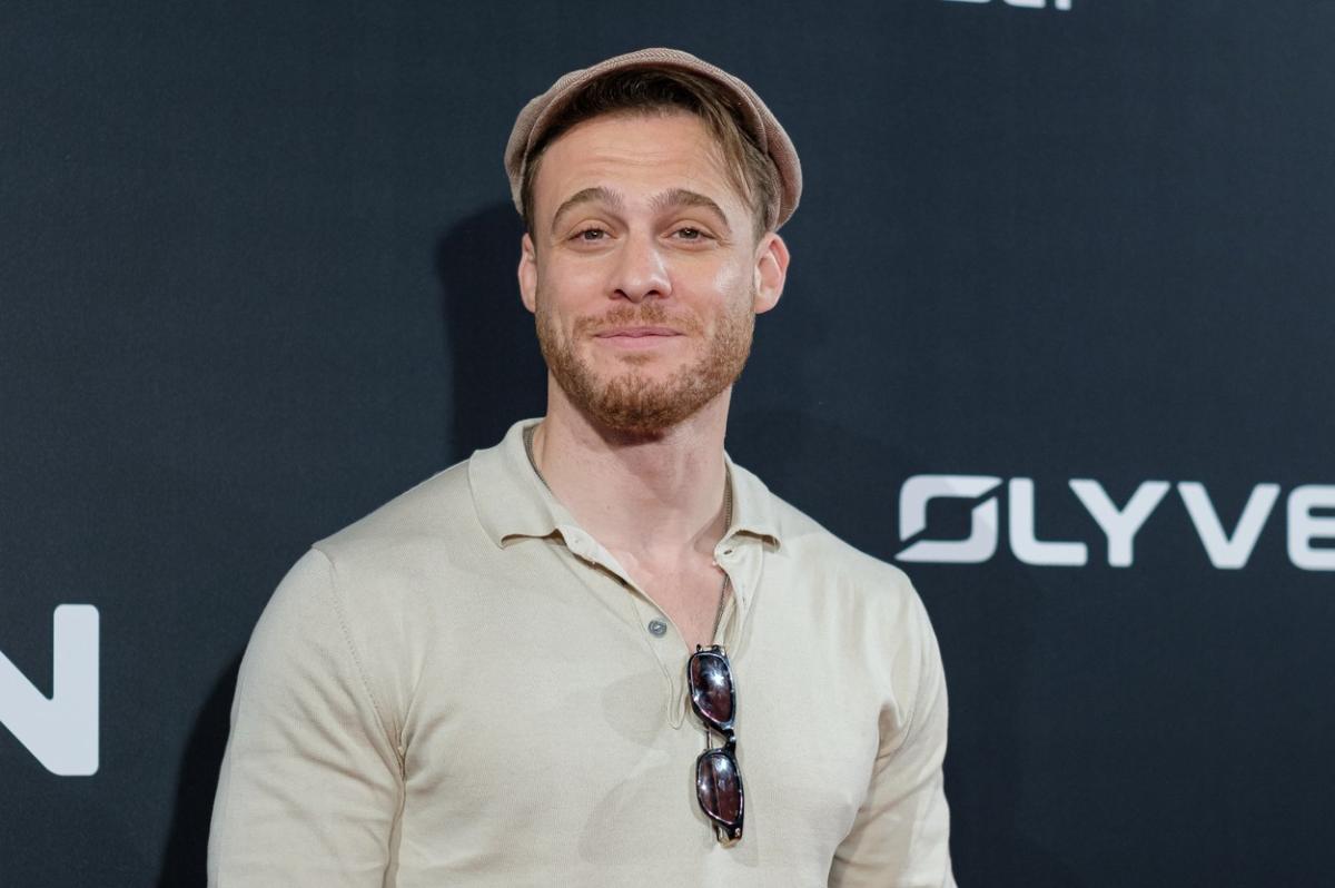 Kerem Bürsin poartă o bluză cu mânecă lungă, chelari de soare în dreptul pieptului și o pălărie