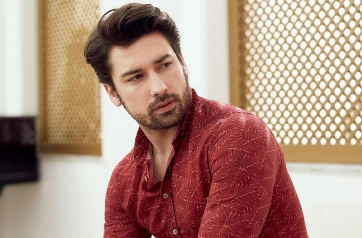 Alp Navruz poartă o bluză vișinie cu mâneci lungi și privește într-o parte