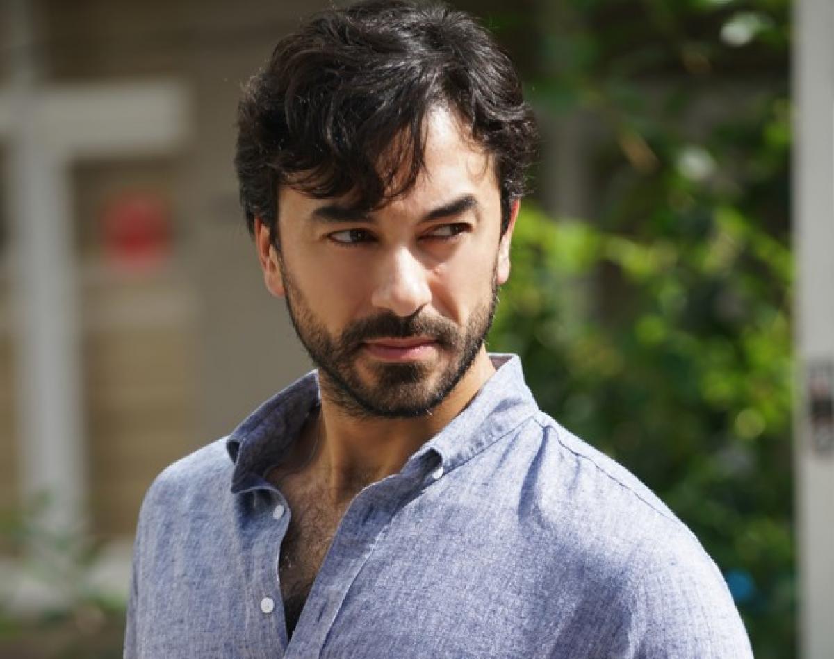 Gökhan Alkan poartă o cămașă albastră cu mâneci lungi, descheiată la piept și are o privire încruntată