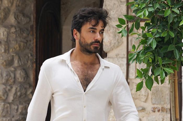 Gökhan Alkan poartă o cămașă albastră cu mâneci lungi, descheiată la piept și are o privire încruntată