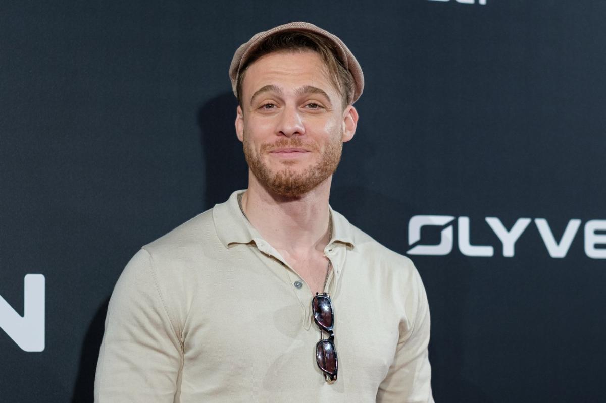 Kerem Bürsin poartă o bluză deschisă la culoare, are ochelari de soare pe care îi ține agățați de tricou și o bască pe cap
