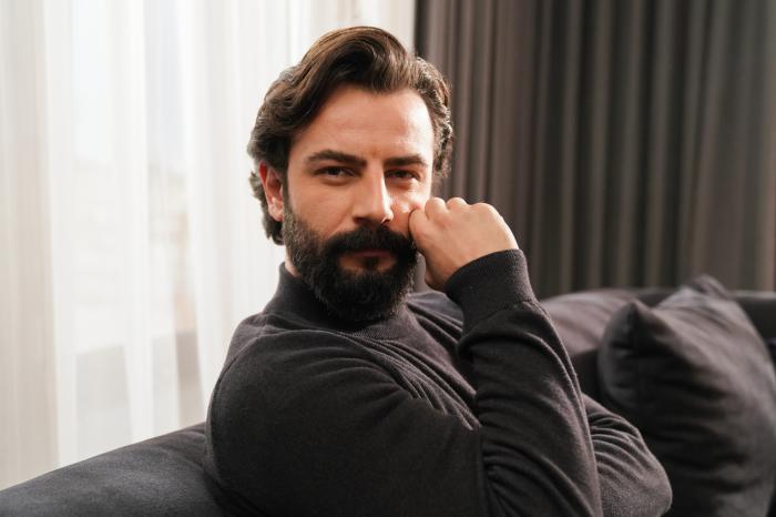 Gökberk Demirci poartă o maletă neagră cu mâneci lungi și ține mâna dreaptă spre față