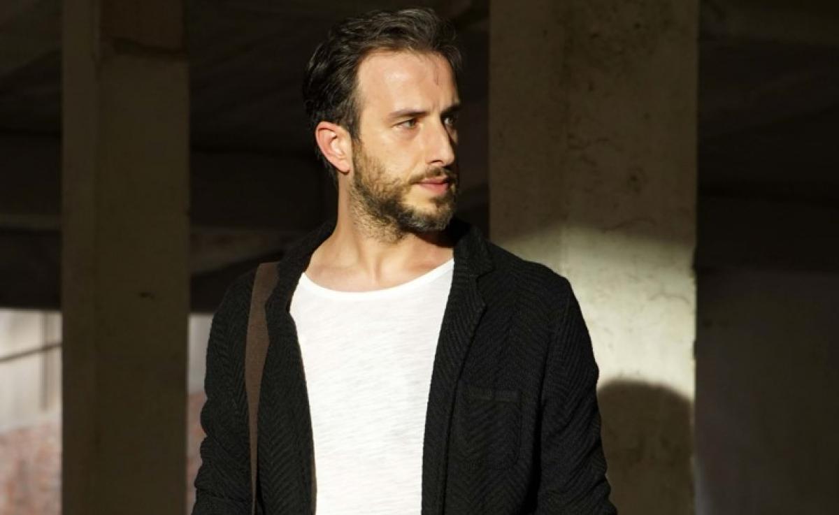 Fatih Artman poartă un triou alb și un sacou negru și ține o geantă pe umărul drept