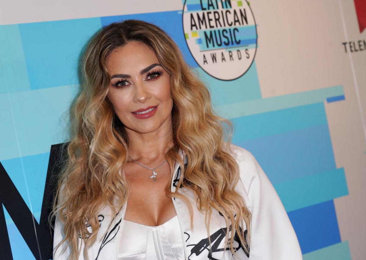 Aracely Arámbula poartă o bluză albă cu decolteu și are părul desprins ondulat