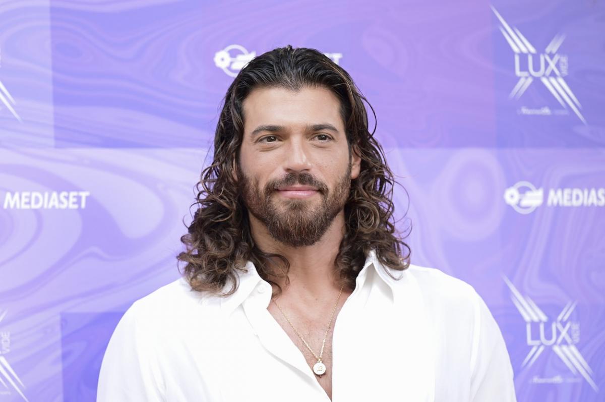 Can Yaman poartă o cămașă albă deschis la gât și are părul lung