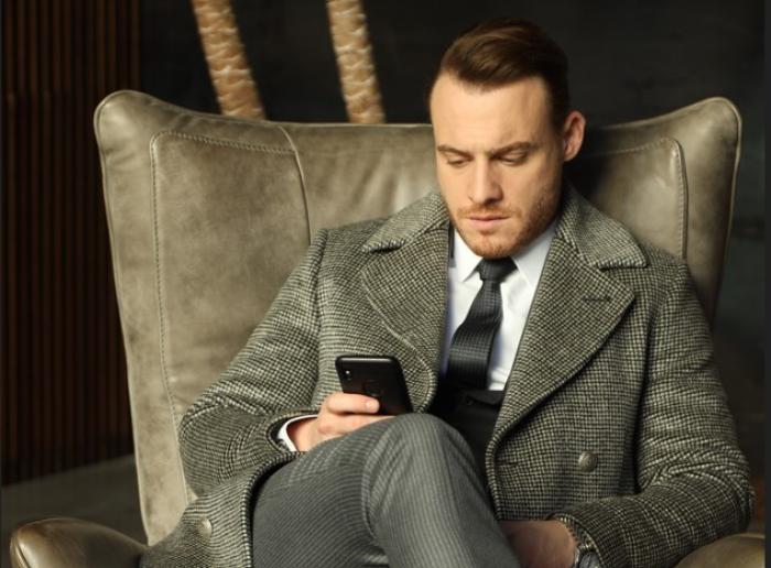 Kerem Bursin poartă un costum, stă pe fotoliu și se uită la telefon