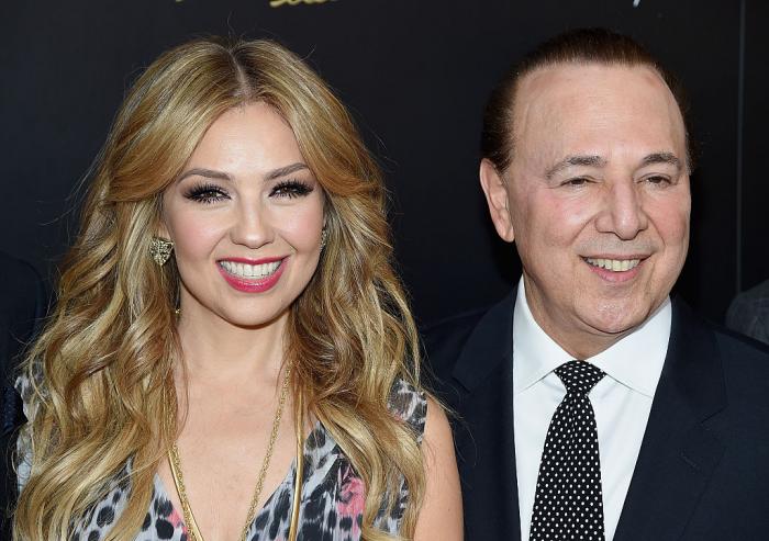 Thalía stă lângă soțul ei, zâmbește și are părul deschis la culoare. Tommy Mottola zâmbește, poartă un costum închis la culoare și o cravată