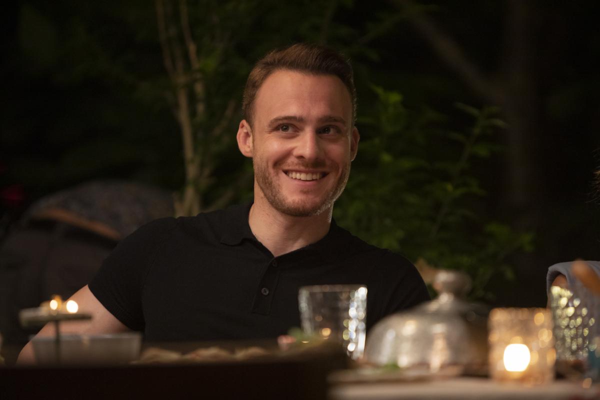 Kerem Bürsin poartă un tricou negru și zâmbește. Actorul se află la masă