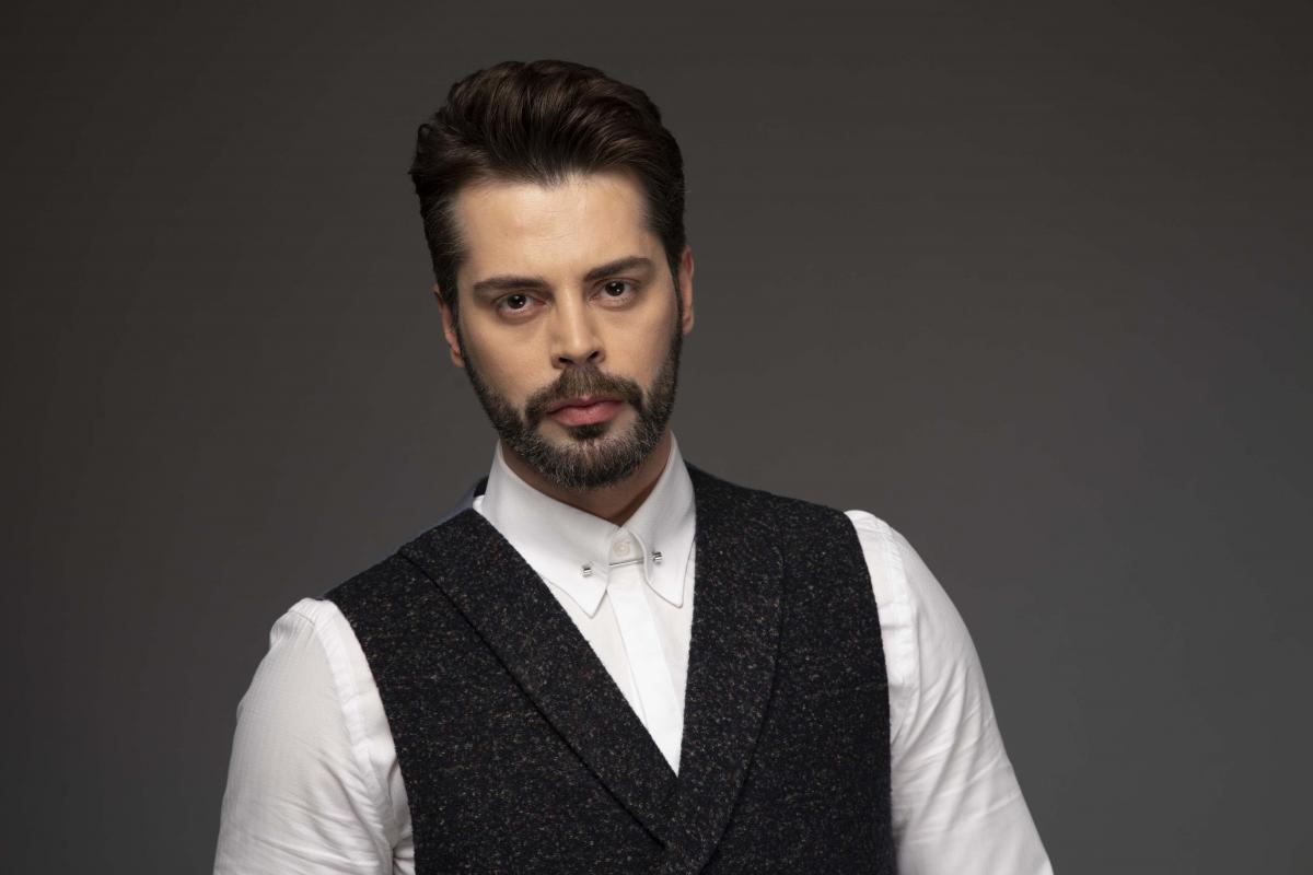 Ali Yasin Özegemen poartă o cămașă albă și un sacou negru. Actorul poartă barbă și mustață