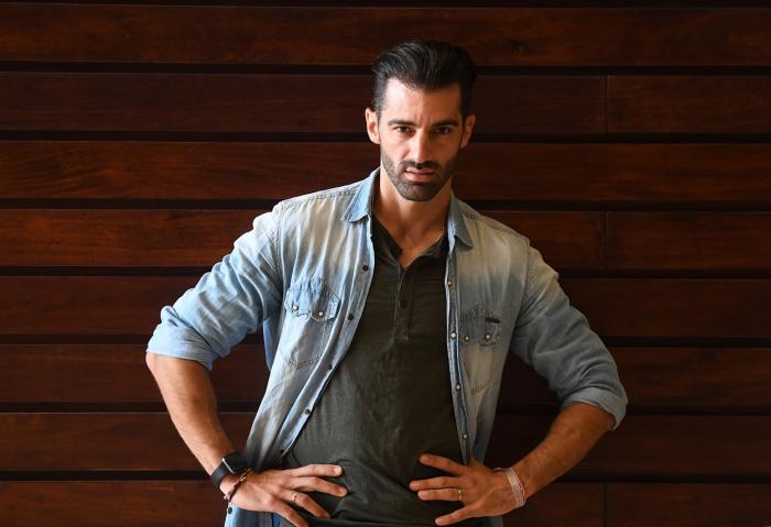 Toni Costa stă cu spatele în dreptul unui perete și își ține mâinile în șold. Actorul poartă o cămașă de blugi