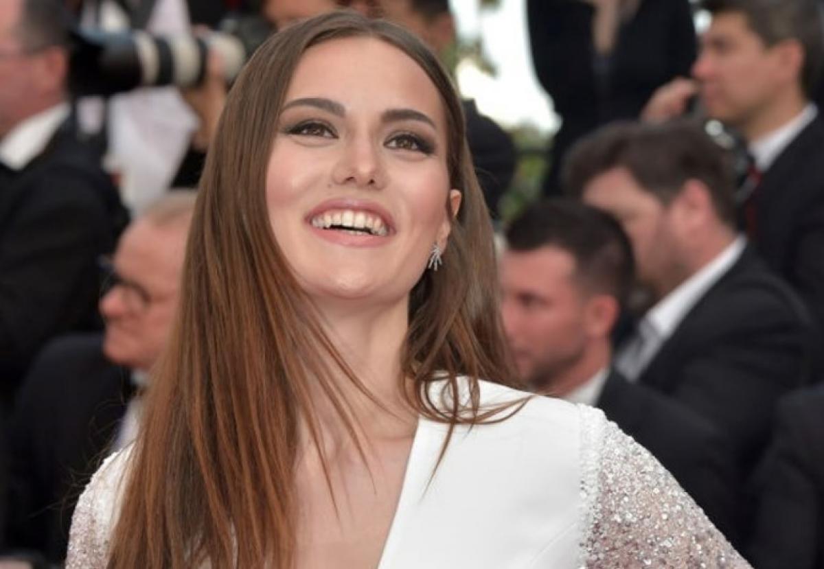 Fahriye Evcen este pe covorul roșu și zâmbește. Actrița poartă o rochie albă și are părul desprins