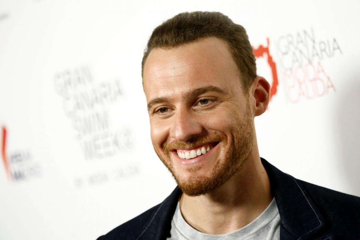 Kerem Bürsin zâmbește în fața camerelor. Actorul poartă un sacou negru și are părul scurt