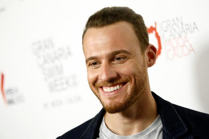 Kerem Bürsin zâmbește în fața camerelor. Actorul poartă un sacou negru și are părul scurt