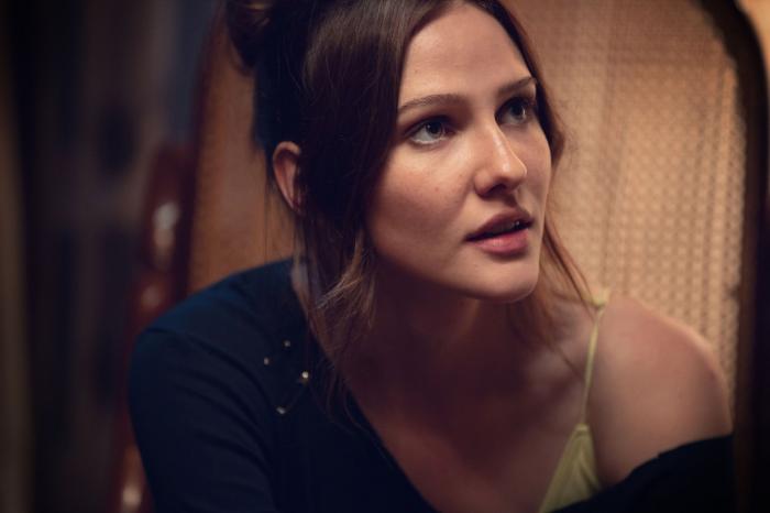 Alina Boz poartă o bluză care îi cade pe umăr, are părul prins sus și breton pe ambele părți
