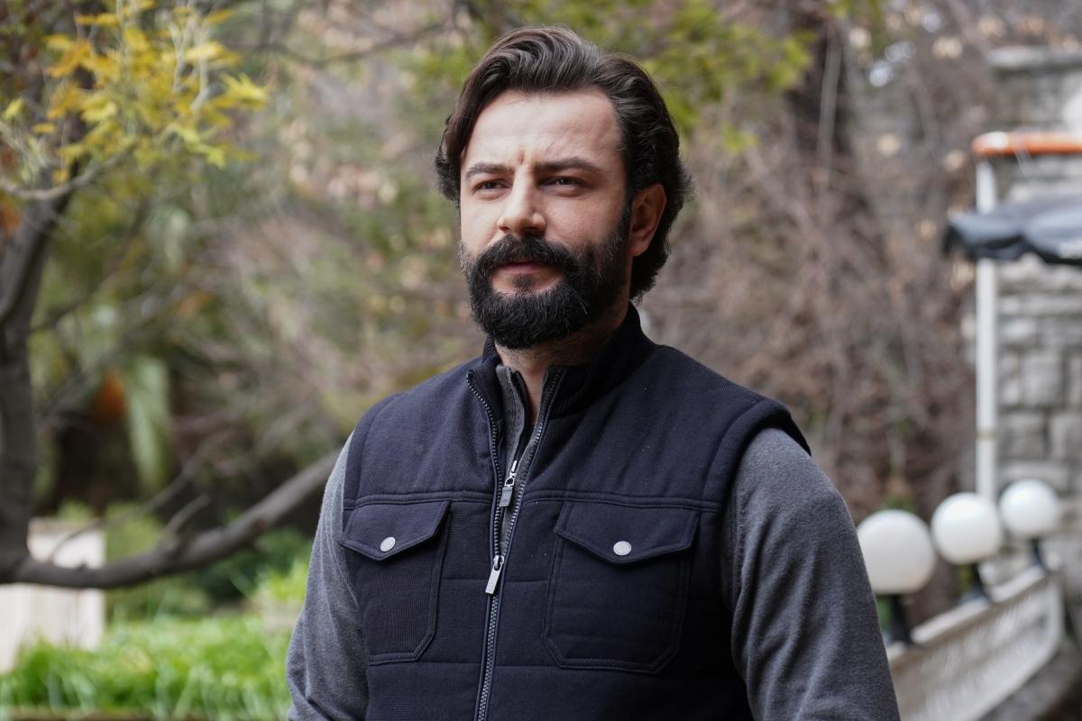 Gökberk Demirci poartă o bluză cu mâneci lungi, închisă la culoare și are o vestă neagră