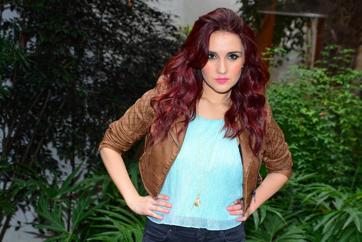 Dulce Maria stă cu mâinile în șold. Are părul roșcat, o bluză albastru deschis și o geacă de piele maro