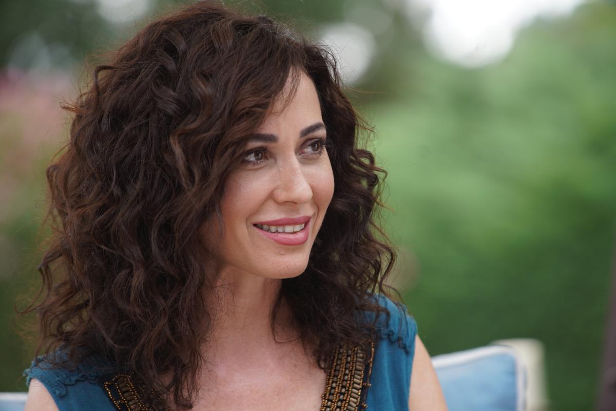 Evrim Doğan zâmbește, poartă o bluză cu mâneci scurte și are părul lung, ondulat, desprins