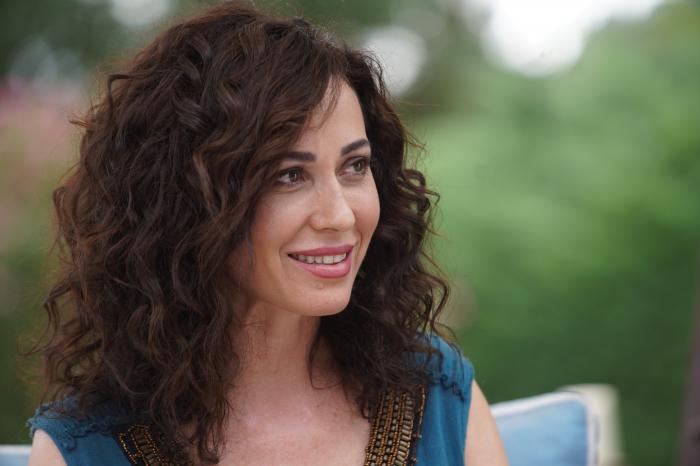 Evrim Doğan zâmbește, poartă o bluză cu mâneci scurte și are părul lung, ondulat, desprins