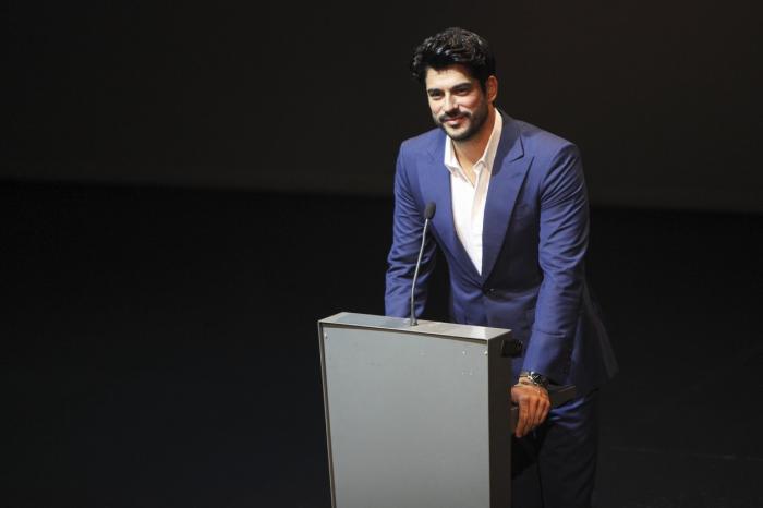 Burak Özçivit poartă o cămașă albă și un costum albastru. Actorul se află pe scenă și ține un discurs
