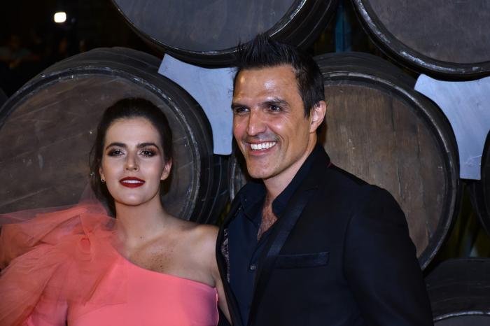 Claudia Álvarez poartă o rochie roz fără mâneci, are ruj pe buze și stă alături de partenerul ei, Billy Rovzar care poartă un costum negru