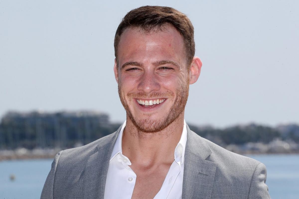 Kerem Bürsin poartă o cămașă albă și un sacou gri și zâmbește