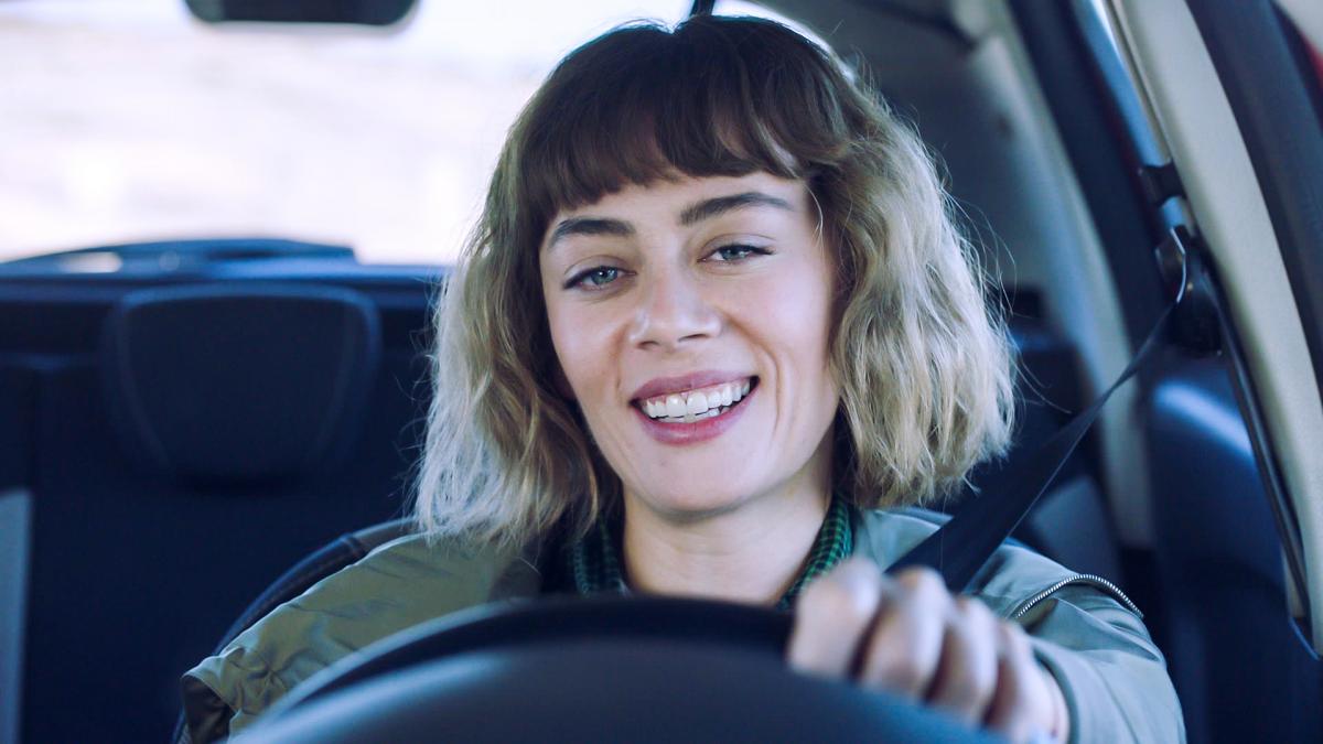Demet Evgar poartă părul scurt, zâmbește și stă cu mâna stângă pe volanul mașinii