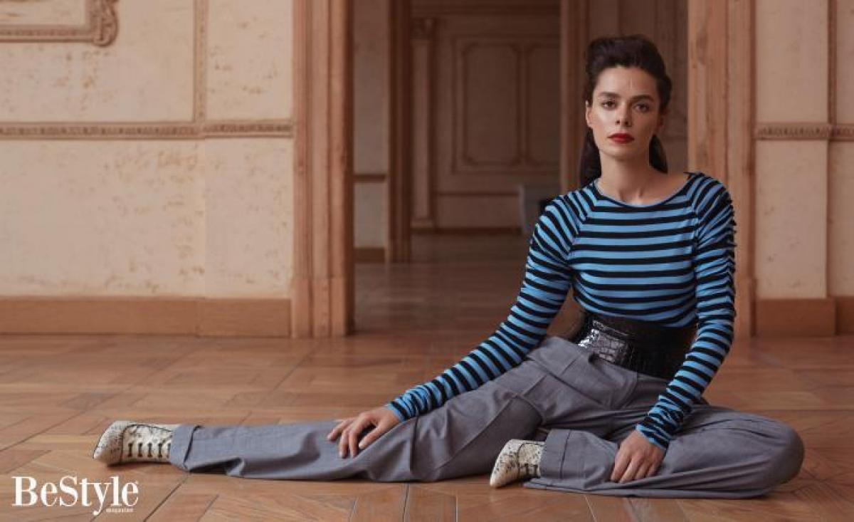 Özge Özpirinççi stă pe podea, este îmbrăcată în pantaloni lungi, bluză albastră în dungi negre și este coafată