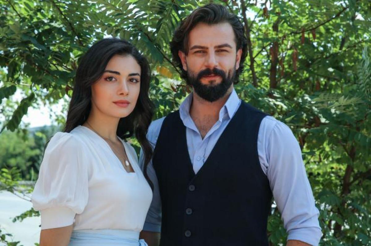 Özge Yağız poartă o rochie albă, părul negru desprins și stă alături de Gokberk Demirci care poartă o cămașă albastră și o vestă neagră