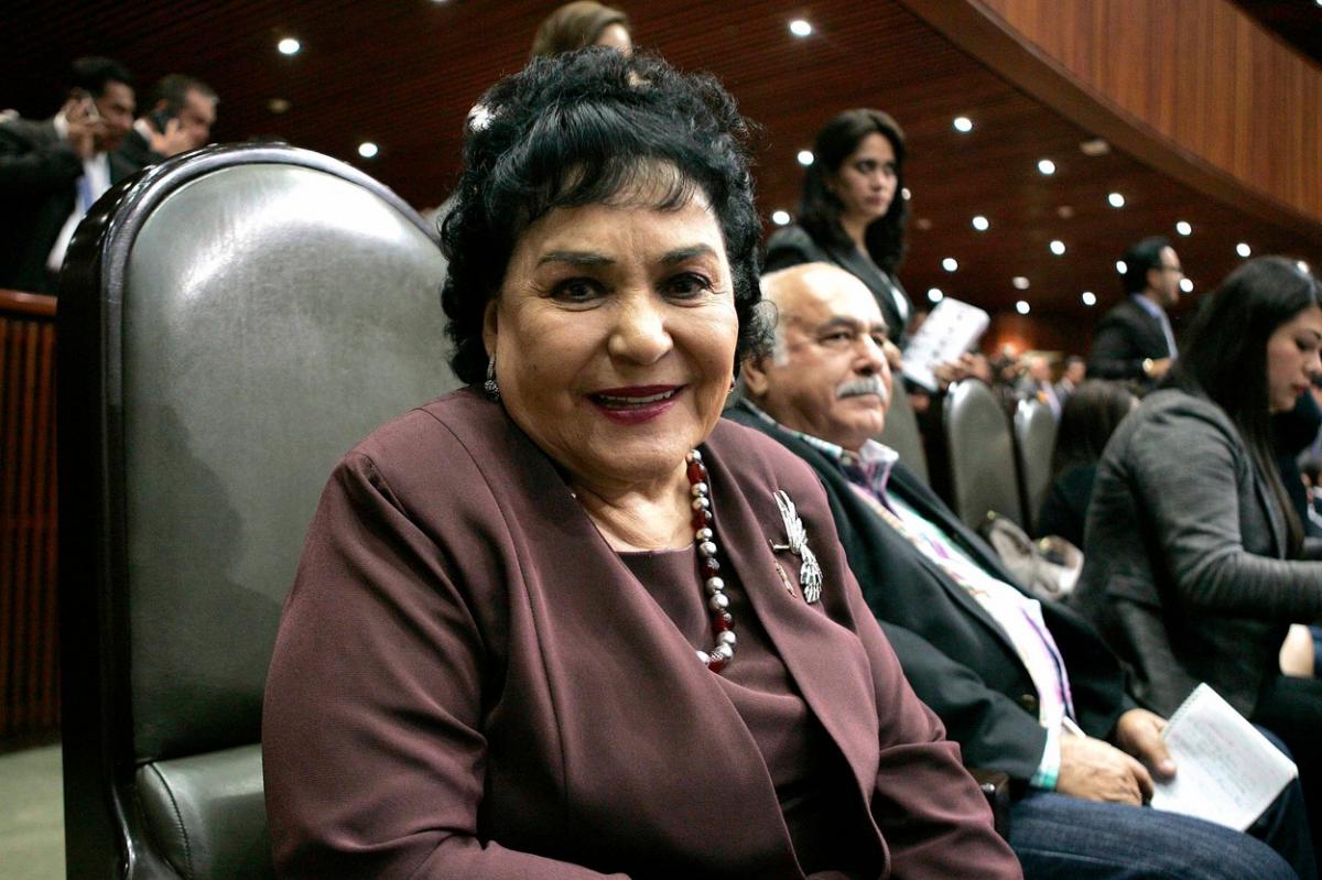 Carmen Salinas poartă un costum închis la culoare, mărgele, zâmbește și are părul brunet